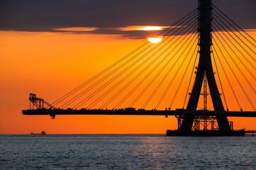Naklejka premium Fiery sunset casting an orange glow over the under-construction Danshui cable bridge in New Taipei Taiwan, creating a dramatic silhouette.