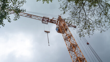 Equipment for the construction and lifting of bulky goods with construction materials. A tower crane on a construction site against a rainy sky. A construction high-rise crane and leaves on a tree.