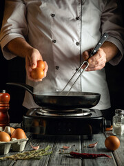 A chef performs meal prep by cracking fresh eggs into a frying pan on a stovetop. The rustic...