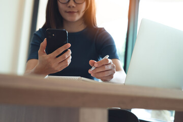 Woman using smartphone and laptop, writing on paper notebook, remote working at home office. Casual...