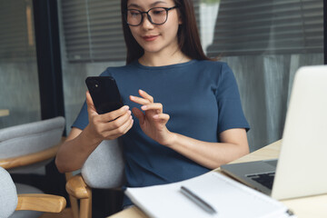 Young asian business woman sitting at table, using smartphone during remote working on laptop...