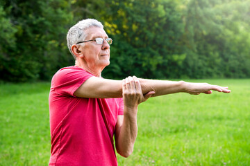 Mature retired sportsman in glasses and sportswear doing side stretching exercises in city park. Healthy lifestyle concept. Copy space.