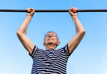 Portrait of an elderly man wearing glasses doing exercises on a horizontal bar at a sports ground. Staying active after retirement.