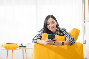 Smiling Woman on Mobile: A young, radiant woman with flowing black hair is engrossed in her smartphone, her face alight with a genuine smile, as she lounges casually on a vibrant yellow sofa.