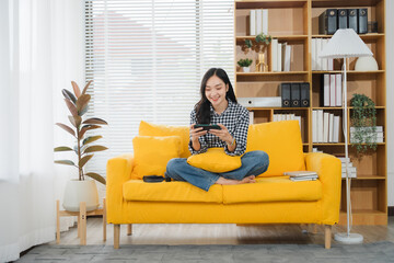 Gaming at Home: A joyful Asian girl enjoys mobile game on a bright yellow sofa, adding a vibrant...