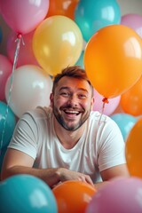A plus-size white man peeks through many balloons, laughing joyfully. The scene is bright and festive, celebrating a birthday party