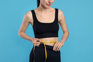 Woman measuring her belly with tape on light blue background, closeup