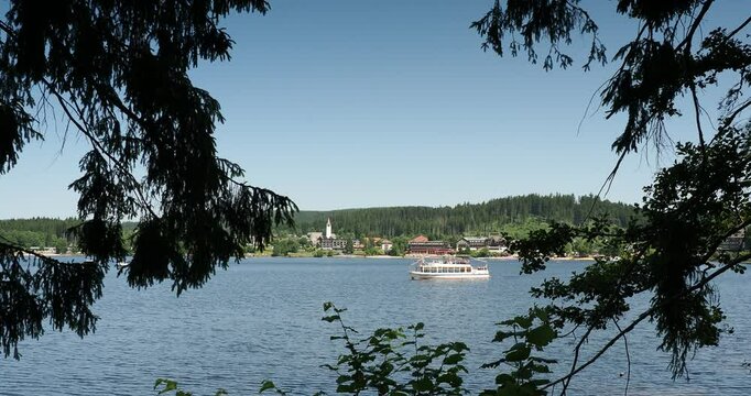 Seelandschaft im S&uuml;dschwarzwald. Kreuzfahrtschiffe auf dem Titisee gegen&uuml;ber der Stadt Titisee-Neustadt vom Seerundweg aus gesehen, im Schatten hoher Buchen und Tannen
