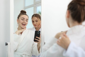 Happy women in bathrobes taking selfie near mirror after spa procedures indoors