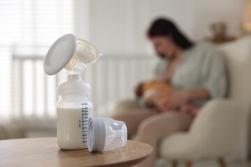 Breast pump with milk on table. Mother breastfeeding her baby at home, selective focus