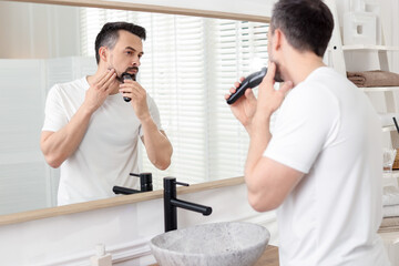 Handsome man shaving with trimmer near mirror at home