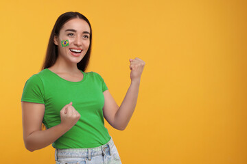 Excited fan with flag of Brazil painted on her cheek against orange background. Space for text