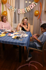 A woman with her son, sister, and nephew celebrates a birthday at a festive table. Soft lighting.