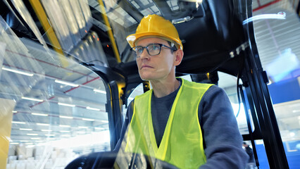 A focused warehouse worker in a yellow safety vest and helmet operates a forklift with precision,...