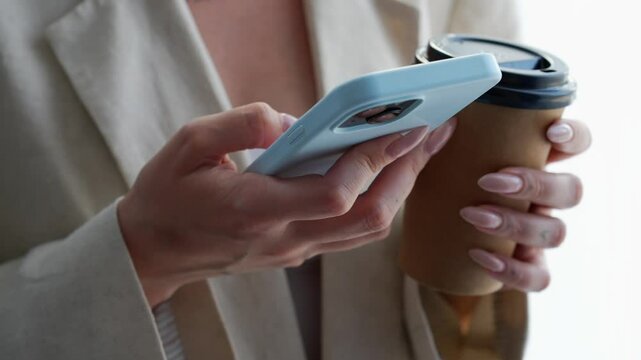 A girl with a coffee glass and a mobile phone in a blue case in her hands stands in a bright office.
The girl clicks the phone and drinks a hot drink from a disposable glass.
Coffee, drink, phone