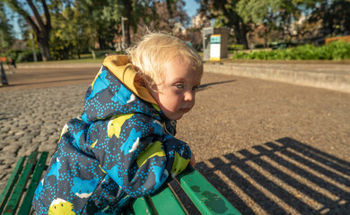 A young blonde child is standing on a park bench.