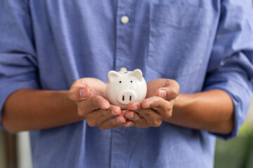 Close-up of man in blue shirt holding ceramic white piggy bank with both hands, representing...