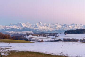 Pathway leading through winter hills on Gurten mountain with colorful evening sky and fameous jungfrau mountain range Eiger, Moench and Jungfrau in the background.