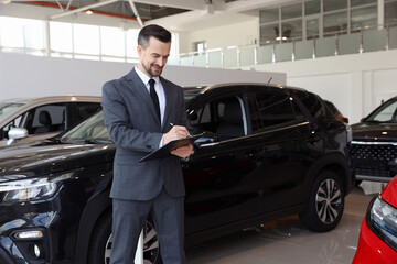 Smiling salesman working with clipboard near new car in salon