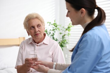 Nurse giving glass of water to senior woman indoors, selective focus. Home health care service