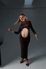 Beautiful pregnant young blonde in a brown dress with an open belly stands against a white wall....