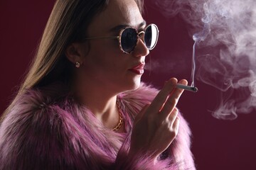 Beautiful woman in fur coat smoking cigarette on dark pink background, closeup