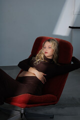 Beautiful pregnant young blonde in a brown dress with an open belly stands against a white wall....
