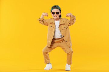 Happy little boy in sunglasses and baseball hat dancing on orange background