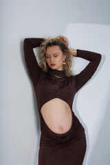 Beautiful pregnant young blonde in a brown dress with an open belly stands against a white wall....