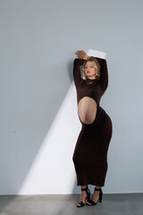 Beautiful pregnant young blonde in a brown dress with an open belly stands against a white wall....