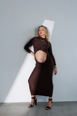 Beautiful pregnant young blonde in a brown dress with an open belly stands against a white wall....