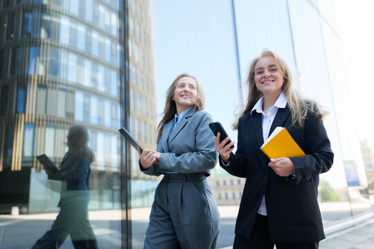 Two professional women walk confidently in business attire, carrying devices amidst modern architecture on a sunny day