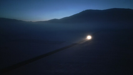 Nighttime aerial view captures a car's headlights illuminating a winding mountain road, symbolizing the journey ahead and the thrill of alpine driving. Darkness and fog add a sense of mystery.