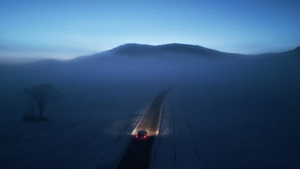 A car travels down a winding mountain road at night, its headlights illuminating the snow-covered...