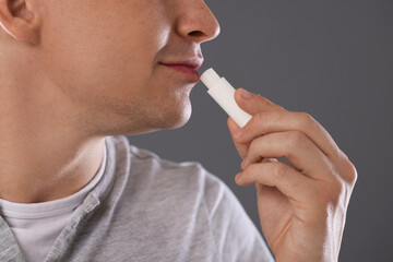 Young man applying lip balm onto his lips on grey background, closeup