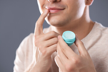 Young man applying lip balm onto his lips on grey background, closeup