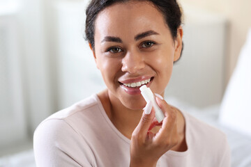 Smiling woman applying lip balm onto her lips on blurred background, closeup