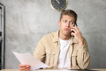 Young man with papers complaining to someone via phone at wooden table indoors
