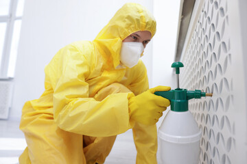 Pest control worker in protective suit spraying insecticide indoors, selective focus