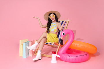 woman in straw hat and heart sunglasses sits on beach chair with arms outstretched with shopping bags isolated on pink background. Perfect for summer promotions.