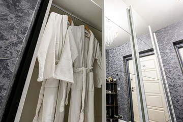 Two white bathrobes hang in a closet with mirrored doors, reflecting a room with patterned...