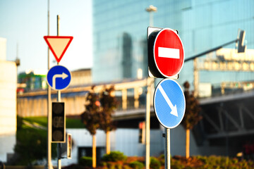 Close-up of multiple traffic signs including no entry, right turn, and give way signs, with modern glass buildings in the background. Concept of road safety, urban transport, direction and navigation  © belavinstock