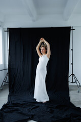 Beautiful pregnant young blonde in a long white silk dress to the floor on a black fabric...