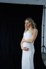 Beautiful pregnant young blonde in a long white silk dress to the floor on a black fabric...
