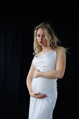 Beautiful pregnant young blonde in a long white silk dress to the floor on a black fabric...