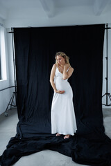 Beautiful pregnant young blonde in a long white silk dress to the floor on a black fabric...