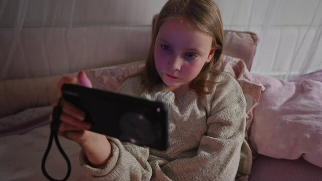 Portrait of Caucasian female child resting on the bed and watching funny video on mobile in her hand. Schoolgirl is spending leisure time online scrolling social media content. Modern technology - Powered by Adobe