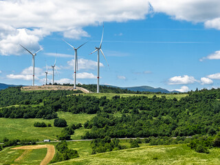 Renewable wind energy turbines standing tall on lush green valley with forest mountains and scattered white clouds