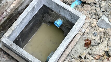 Unfinished concrete drain pit at messy construction site filled with murky water. blue PVC pipe for new sewer system reveals challenging plumbing work underground