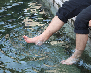 Man cooling his feet in fresh water, sitting on the edge of a stone wall. Summer relaxation and nature therapy concept.